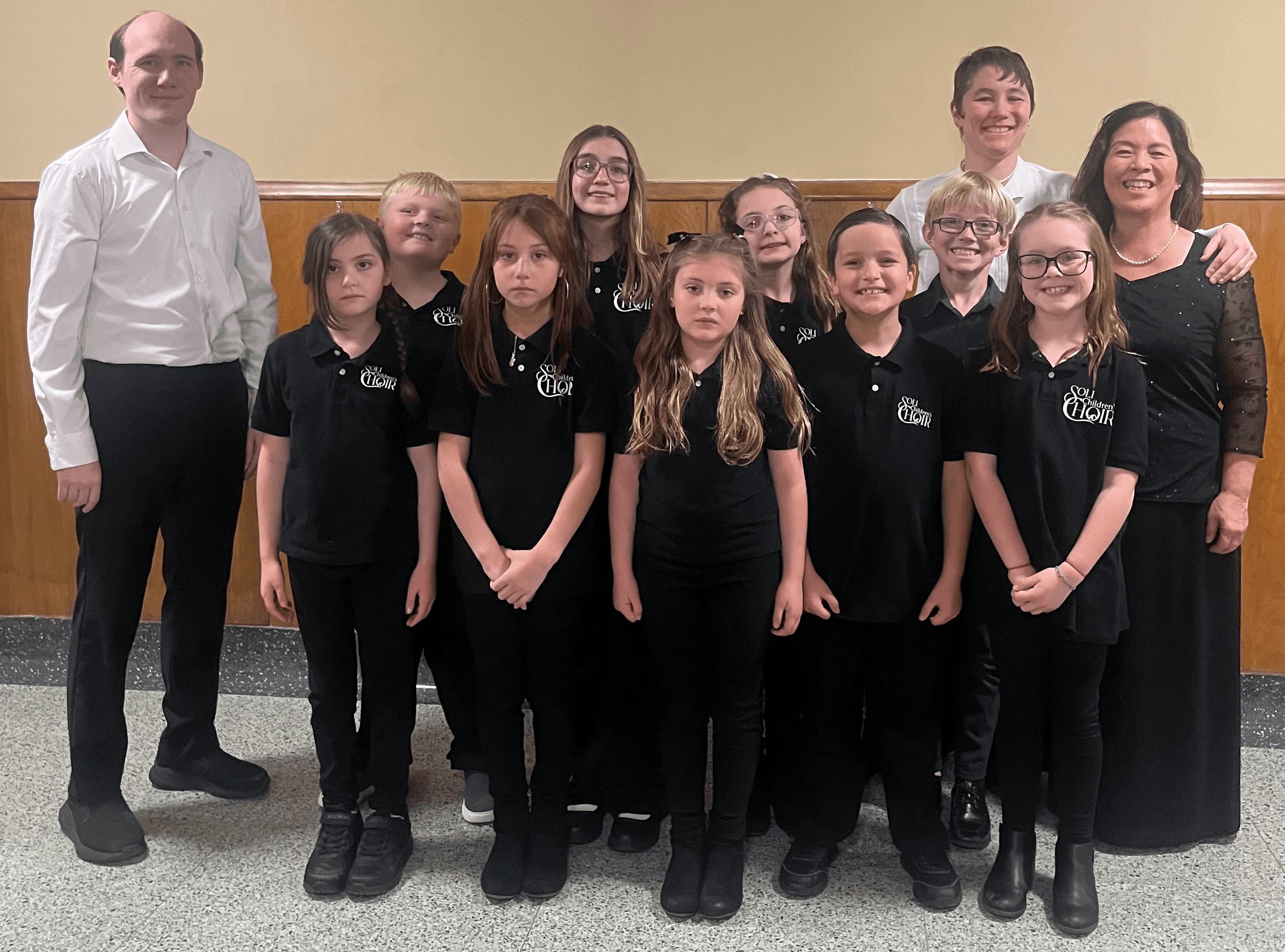 Members of the Soli Children's Choir in black choir polos, standing with three adults