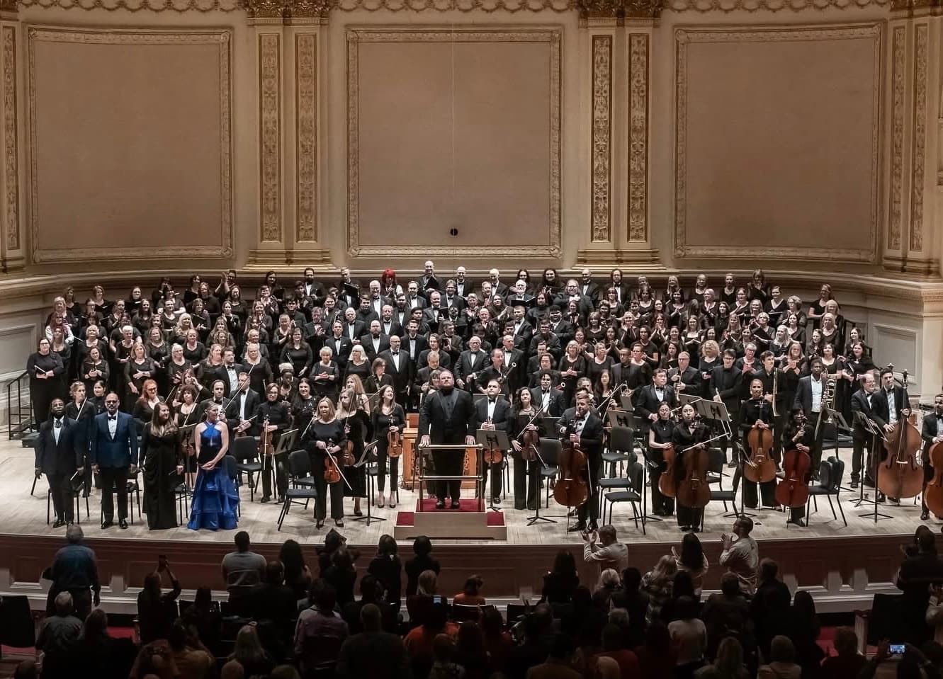 The Concert Choir and orchestra on the Carnegie Hall stage, the conductor at the podium, as the audience applauds